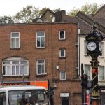 A metal clock on a pole. The pole is black, with red shields at the base and most of the way up, and gold decorative detailing, including crosses on the upper shields. The clock is four-sided, with a triangular pediment (with gold detailing) above a white clock (with black hands) on each face. Above that is a spire, with a black base and gold point. Behind are many shops, brick on the left and white/blue render on the right. A lorry passing by can be seen, just, at the bottom.