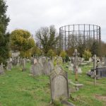 A field of headstones, with grass between. On the right and in the background are trees, some losing their leaves. On the skyline, behind the trees, is an empty gasholder.