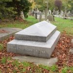 A tomb. It is of two tiers, grey marble. The upper tier is set back from the lower, with sloping sides joining the two. The upper tier has a cap shaped like the hipped roof of a house. The longer side of the top has writing (illegible, from this angle); the short (front) side of the lower tier reads “CHARLES BABBAGE, ESQ.” Behind are many other tomb stones, mostly old but a few new, and some trees.