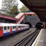A Tube station platform. The station is open-air but in a cutting; there is a train at the far platform blocking the view of it. The near platform has a few people on it, on sitting and the rest standing. There is a footbridge with steps leading up, red-painted, from the canopy, with the bridge passing over a tunnel mouth. The tunnel mouth is at the far end of the station, and has two circular portals. Above the far platform, there are a few trees and there is a row of buildings.