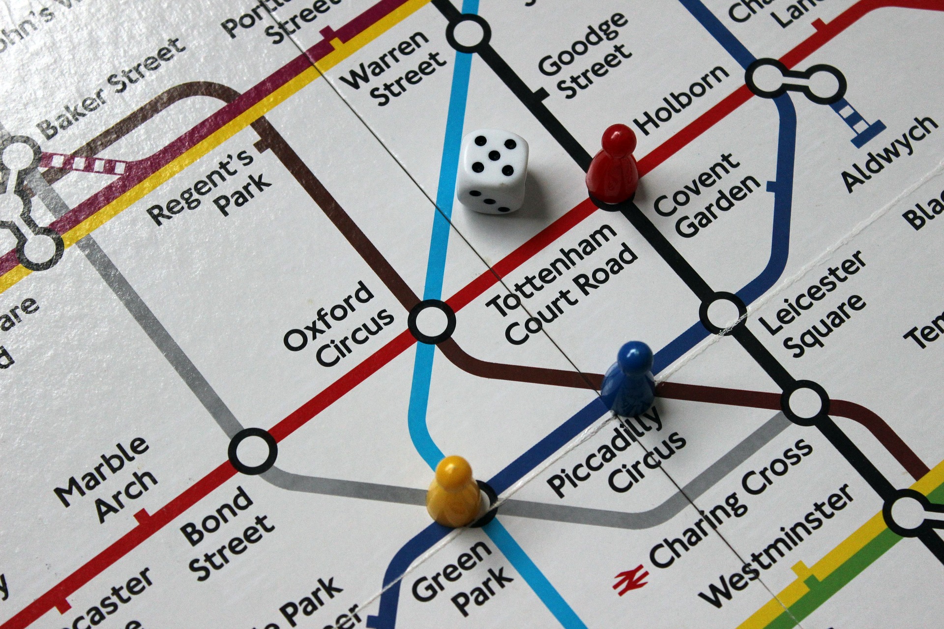 Three pawns (red, blue and yellow) and a die on a game board printed with the Tube map. The central section is visible. The die is in the triangle formed by the Victoria, Northern and Central lines, and the pawns are on Holborn, Piccadilly Circus and Green Park (red, blue and yellow, respectively).