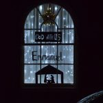 A house window, square at the bottom and arched at the top, divided into panes. The house is dark; only the light from the window, and its reflection in a car below, can be seen. The window has white fairy lights in front of white curtains; on the window are black silhouettes of a black rectangle (with “God with us”) cut out, the word “Emmanuel”, and Mary, Joseph and a manger in a stable. Above all the silhouettes is a gold star.