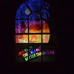 A window, square at the bottom and arched at the top, divided into twelve panes. The surrounding is dark, so only the light through the window can be seen. The window has a translucent design on it, of a dark sky (with moon) and silhouetted tree, and two people and a deer around a campfire. Beneath the whole is a darkened part, with writing in different colours with light shining through: “Love keeps the cold out better than a cloak”.