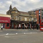 Across a road, a symmetrical yellow-brick pub, with bay windows on either side. The pub is “The Old Bel”. To the right is a red-fronted amusement arcade, that looks closed, and to the right a modern block of flats. There is a number 32 red bus leaving the picture to the right, and there are various pedestrians on the pavement.