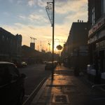 Looking down a street with shops on either side, in modern looking buildings silhouetted against the sunset. There is a hanging basket, suspended from a lamppost, in front of the setting sun; the sky is a range of blues with pink clouds. Also on the lamppost is a shooting-stars Christmas decoration.