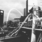 An arches bridge over a railway station, in front and to the right of the arch of Wembley stadium, with various high-rise buildings surrounding. The photo is heavily-processed and in black-and-white.