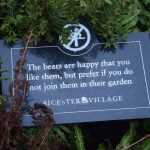A black marble sign in foliage. The sign has, in white, a pictogram of someone climbing on a ledge, crossed out, and below it, “The bears are happy that you like them, but prefer if you do not join them in their garden”; followed by “BICESTER VILLAGE”, with a rose logo between the two words of the site’s name.