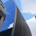 A long and tall building. The end wall is glazed, and egg-shaped; the side wall, following the contours of the egg, has narrowly-spaced metal ribs. Other buildings surround.