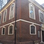 A corner view of a large two-storey brick building; the upper storey has high arched windows (five on the left, three on the right); the lower has lower arched windows (five on the left, two on the right either side of an ornate door). The windows have white frames. There is a plaque next to the door, and there is a bench by the door. Three old-fashioned lamps hang from the buildings. The surrounding buildings are mostly more modern.