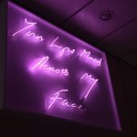 A pink neon sign in a dark space, below an artificial roof with an unlit spotlight. The pink neon light is handwriting, reading “Your Lips Moved Across My Face”.