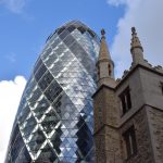 The Gherkin, behind a square yellow-stone church tower with four small spires at the corners. An office block is to the left. The sky is blue with white clouds.