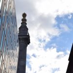 The Monument to the Great Fire of London, looking up against a partly-cloudy sky. There are two office buildings in front.