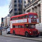 A road junction, with office buildings along the road leading away and a House of Fraser department store, with Ionic columns along its facade, on the right. In front is traffic, including a red Routemaster bus: “15: St Paul’s Cathedral, Fleet Street, Aldwych, TOWER HILL”. The bus carries adverts for Mamma Mia! at the Novello Theatre.