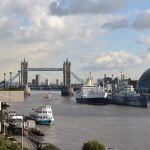 Looking down the Thames from London Bridge; Tower Bridge can be seen. In the Thames is anchored a modern white ship next to HMS Belfast. The sky is ominously cloudy.