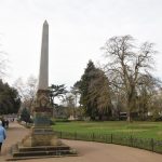 A grey-stone obelisk on a yellow-stone plinth, with bronze plaques attached, covered in verdigris. A beige path snakes off to the left, while there are formal ornamental gardens to the right, separated from the path by a low fence. Trees of many types are around.