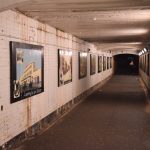 A tunnel, with a tarmac floor, whitewashed brick walls and a whitewashed ceiling. Along each wall are painted images of the town. The tunnel is lit by fluorescent lights. At the end of the tunnel, in the dark opening, a woman with blonde hair is standing.
