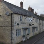A street of Cotswold stone old house, with slate roofs; the nearest one in the terrace in the Carpenters Arms pub, with leaded windows and two doors with stone porches. The pub has a satellite dish on its near wall.