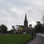 A grasses area, taken from a path along its right-hand side; the path is separated from a road by a low white fence, with cars along the road. A family play football on the bass, while some other people walk dogs. A stone church, with a central tower and spire, are at the far end, and other buildings across the grass. There are trees on the right-hand side.