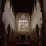 A view of a church from behind the altar, looking towards the stained-glass west window. The roof is of dark wood, and there are clerestory windows in white walls above stone arches.
