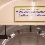 In the foreground, on a high wall, a sign saying “District and Circle lines / Westbound platform 1 / Eastbound platform 2” with a forward arrow; there are green and yellow stripes at the top of the sign, with the text in blue on white. Behind can be seen the beginning of the ceiling of the escalator shaft, with a hanging sign that says “JUBILEE LINE / NORTHERN LINE / BRITISH RAIL”.