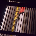 Looking down through a grille in the floor to a Tube train (white, with a red door) at a platform. The raised dots at the edge of the platform can be seen, along with a yellow line and, at the very edge, the words “MIND THE GA”, the last letter being obscured.