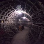 A curving undecorated tunnel, lit by strip lights on the ceiling. The walls are divided into segments. Various detritus is to the left of the paths, including sections of rail.