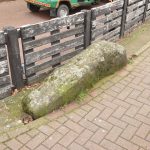 A lumpy grey rock, set into the ground behind herringbone brick tiling. It is in front of a wooden fence; painted brown (the paint is peeling). A red forecourt is behind, with part of a teal-coloured vehicle visible.