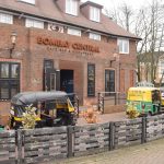 Bombay Central Café Bar & Restaurant, a brick building with a red roof, a former pub. There is the top of a red lion rampant above the porch. In the forecourt are various little vehicles. In front is a little fence, with a stone embedded in the ground near it.