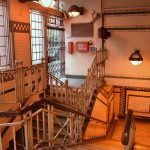 Criss-crosing stairways leading down into a Tube station from ground level, where the photo was taken (at the top of one of the stairways. There are iron railings with wooden handrails, and tiled walls with simple geometric patterning. The windows out onto the street are foggy. At the top of the other staircase is a gated-off door.