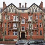 A brick apartment building, with mock-Tudor detailing on the gable ends. The apartments have balconies, and some arched windows. In front is a row of coppiced trees. There are cars parked on the road outside.