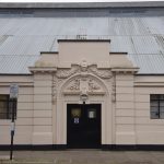An archway, painted cream, over dark double doors. The archway has intricate decoration, including garlands and a head, above the door, and two columns either side. To either side of the door are plainer walls, with windows set in them. Above the door is a high corrugated metal roof.