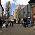 A pedestrianised shopping street, that curves around to the left. There are several shoppers, and a few trees whose leaves are just starting to appear. The most prominent shop is a four-storey art-deco building with cream tiles, which is a House of Fraser Outlet Store; there is a white sign in every window saying "Closing Down, 20% Off".