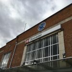 A brick art deco station frontage, viewed from below. There is a glass canopy at floor height, with high windows above, and above that the sign reading "Doncaster". Above that is a large blue clock with silver hands and numerals. The sky is angry.