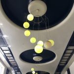 A long ceiling, stretching away from the camera. The ceiling is white, with blue ornaments: two strips divided into squares along either side, and three large circles. The circles have white circles inside them, from which hang light fittings made of blue orbs, descending in a helix (supported by wires).