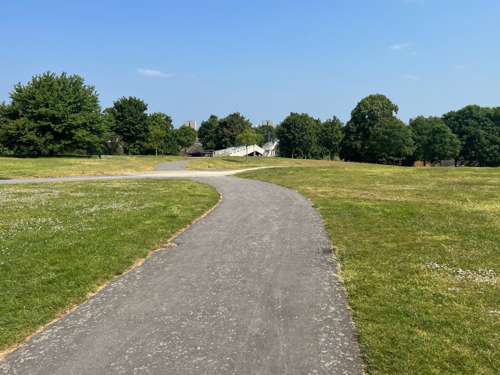 A view along a tarmac path in a park. The path crosses another in the middle-distance, and between them all is green-brown grass speckled with daisies. The horizon is formed of a row of trees with dark green leaves, puncutated only by a concrete footbridge, and with two tower blocks protruding from above them in the middle. The sky is blue with a couple of tiny white clouds.