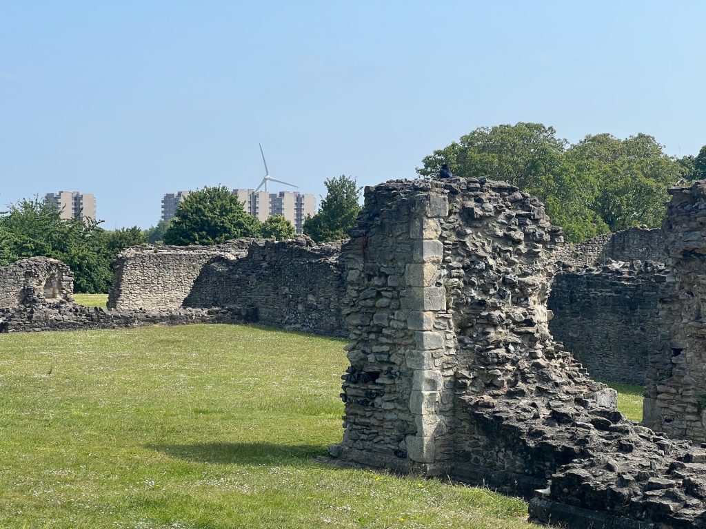 Some abbbey ruins in grass beneath a blue sky. The walls only extend a few feet above the gorund, and the stonework is very rough even where it hasn't been destroyed. In the back left, behind a line of trees, are some tower blocks and a wind turbine.