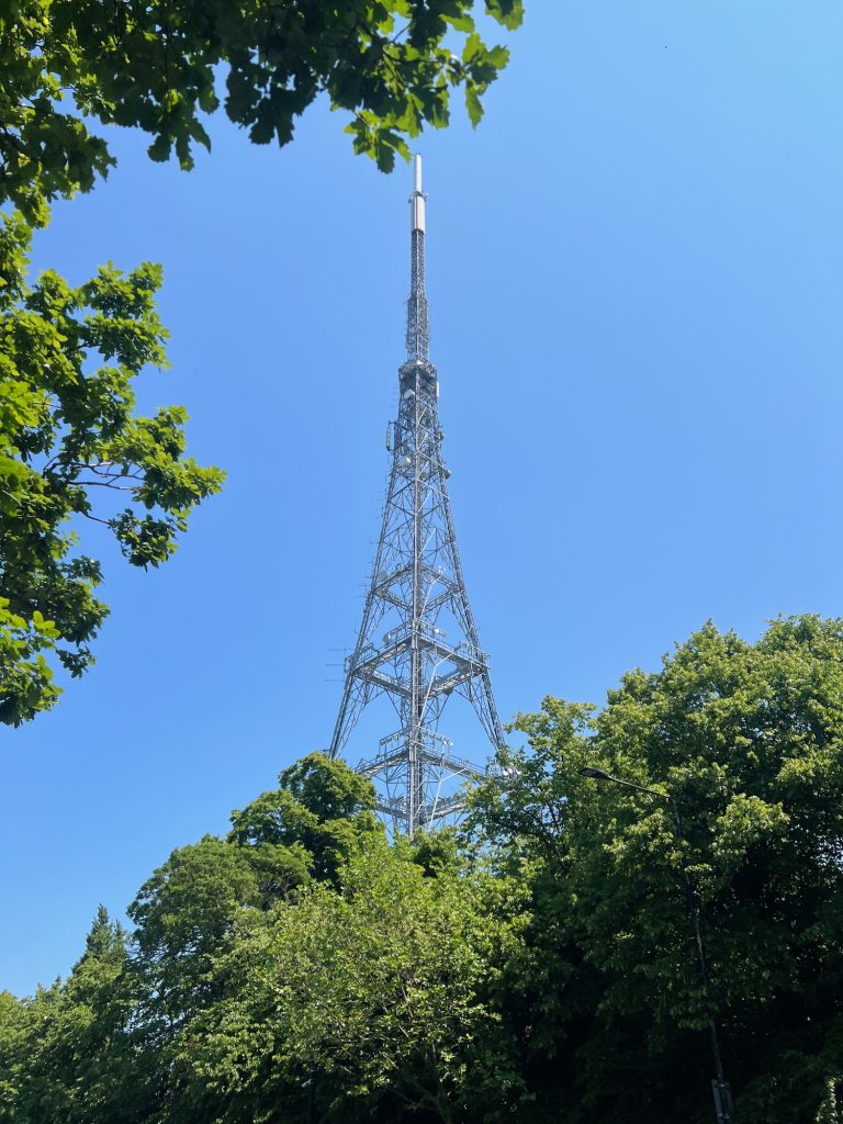 A lattice transmitter mast, viewed from near the bottom looking up to the top, as it tapers to a point. It seems to rise up from above leafy trees, with others overhanging on the left. The sky is bright blue.