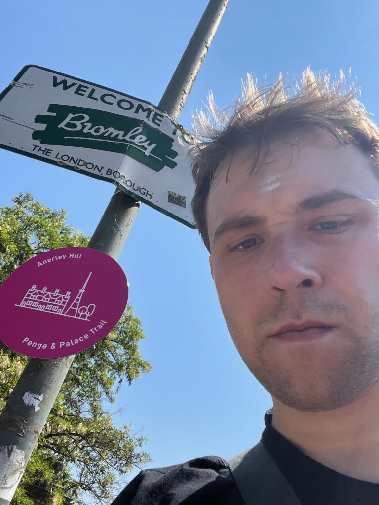 A selfie of the blog's author, peering in from the bottom-right of the photo with sweat shining on his forehead. To the left is a lamppost with two signs, the first reading "WELCOME TO Bromley, THE LONDON BOROUGH"; it is rectangular, with green text on a white background with a green border (except for the name of the borough, which is written in cursive writing in white on a rough green patch). The second sign, smaller and lower down, is circular and pink, with white text around the edge reading "Anerley Hill – Penge & Palace Trail"; surrounding a minimalist line drawing of a large house, a transmitter, and two trees. A tree pokes in behind the sign, and the sky, which forms most of the background, is blue from top to bottom.