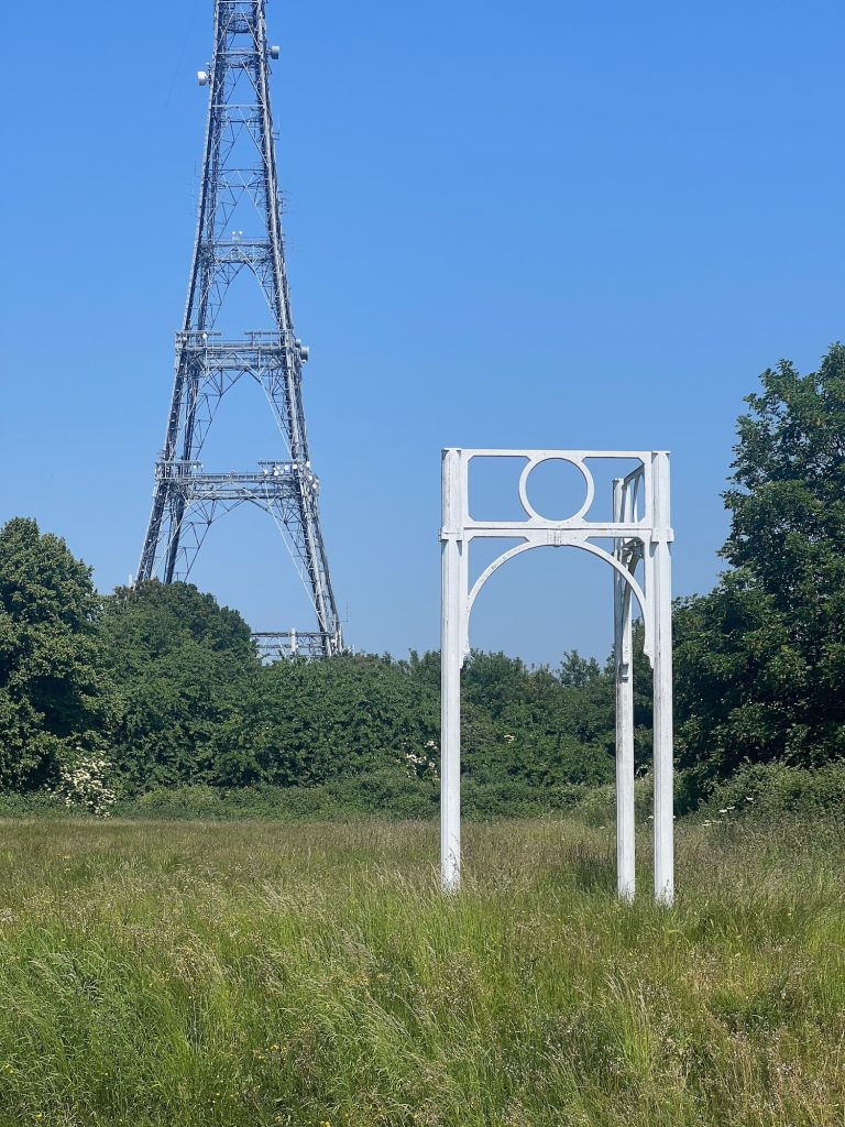 A sculpture in a park in the shape of two ironwork arches, set at right-angles, each topped with a rectangle above with a circle of iron inside. It sits in rough grass in a park. In the background, against a solid blue sky, a latticework tower rises up from some leafy green trees.