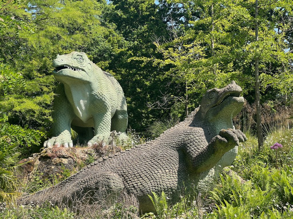 Two sculptures of dinoaurs. The one further back is slightly higher; it is light green, standing on all-fours, and facing the camera. Below it is a brown dinosaur, facing right but with its head turned towards the camera, raising its right foreleg. They sit in the bright sunlight among trees and shrubs typical of a park.