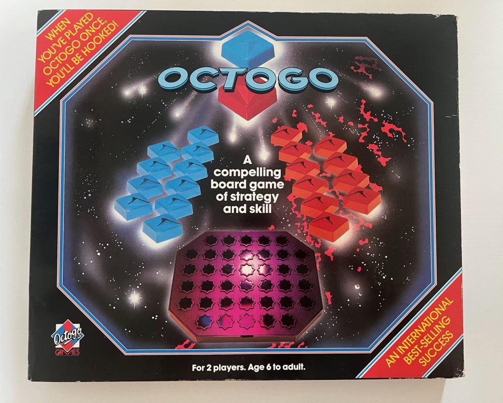 A box for a board game called "Octogo". The box is black, with an octagonal main image showing a space-themed background. At the top of the image is the game's name, above two of the playing pieces, plastic squares with arrows on them, the top one blue and the bottom one red. (The border of the octagon distorts around the square.) Below it ten pieces of each colour (blue on the left) are shown facing each other, with the text "A compelling board game of strategy and skill" in white between them. The game board, which is a plastic octagon with 38 eight-pointed stars on it, is shown at the bottom. Below the image is the text "For 2 players. Age 6 to adults." Red stripes cross the top-left and bottom-right corners, with yellow text on each; the first says "WHEN YOU'VE PLAYED OCTOGO ONCE, YOU'LL BE HOOKED!" and the second says "AN INTERNATIONAL BEST-SELLING SUCCESS". The bottom-left corner shows the logo of Octogo Games.