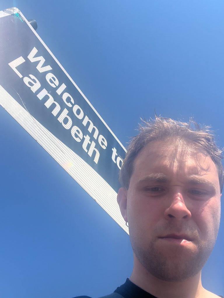 The blog's author, looking down at the camera with a scowl on his face. His hair is messy, and appears fairly short because of the camera angle. Behind him is a sign. The sign is dark blue above a curved line, and white below it (the dark blue area being much bigger than the white), with a white border. On the dark blue area "Welcome to Lambeth" is written in white. The sign is visibly coated with a retroreflective material. Behind the sign is a cloudless sky, paler at the top-right than at the bottom-left.