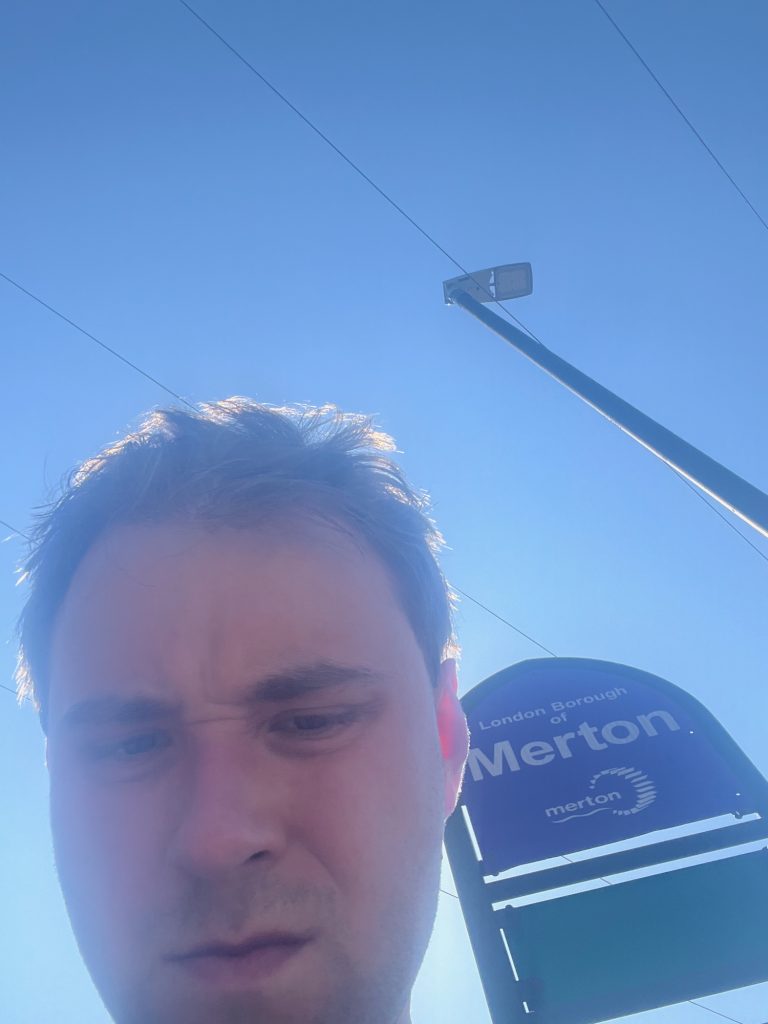 The head of the blog's author, very close to the camera. Behind him is a sign supported by a metal tube shaped into a pointed arch. The upper part of the arch holds a purple sign reading "London Borough of Merton", above the Merton Council logo (the word "merton", in lowercase, with a wavy line beneath, intersecting a waterwheel), all text and imagery beind in white. Below it, and separated by a metal pole, is a blank green sign. The camera angle is noticeably upwards. To the right of the sign is a lamppost, and three telephone wires cross the sky, which is cloudless (the sun appears to be behind the man's head).