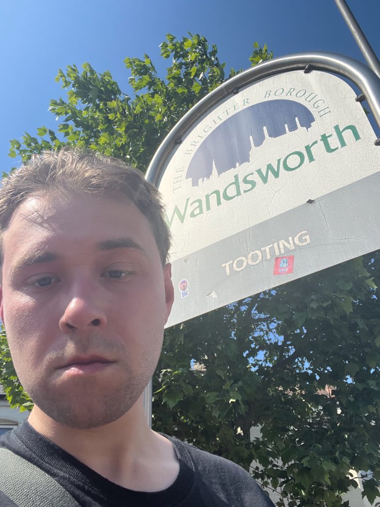 The blog's author in front of a sign supported by a metal arch. The sign inside the frame is likewise arch-shaped, and white, with a grey bar along the bottom. On the white part is the word "Wandsworth" in green, beneath a blue semicircle, the bottom edge of which is a skyline including a church tower and Battersea Power Station. Arching above the semicircle, text in green says "THE BRIGHTER BOROUGH". Below, "TOOTING" is written in white text on the grey bar. Behind the sign is foliage, with a building behind that which is hard to discern through the leaves. The sky is solid blue.