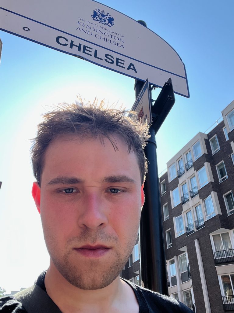 The blog's author, squinting at the sign, his hair messy and his ears turned bright pink by the sunlight streaming through them. He stands between the two poles of an arch-shaped white sign, which is divided into two parts by a blue line. Above the line, in blue, is a coat of arms, above the words "THE ROYAL BOROUGH OF KENSINGTON AND CHELSEA". Below the line, in black, is the word "CHELSEA"; there is also a sticker on this part of the sign. There are two smaller signs on the right-hand pole, not clearly visible. In the background is a modernist block of flats, made of dark brick and render, with windows arranged irregularly. The sky is bright blue, with the sun forming a sort of halo around the man's head.