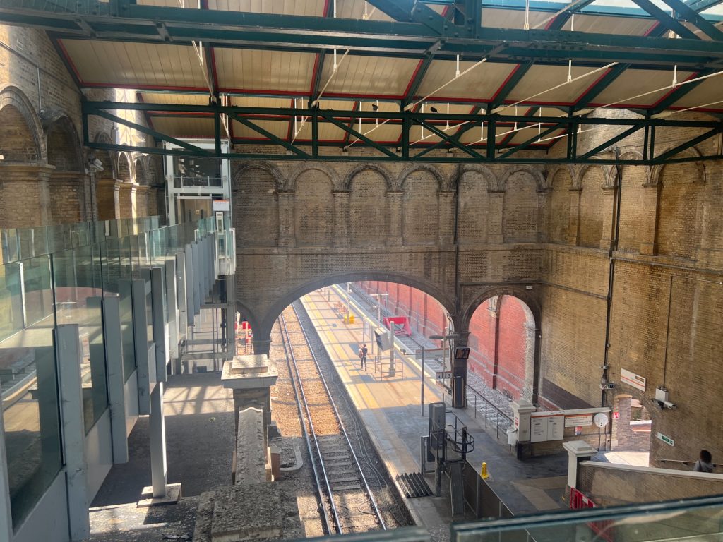 Looking down on a large yellow brick train shed from a bridge. On the far wall, there are three arches: two narrow ones on either side flanking a large one in the middle. A wide arch spans a track and a platform, while the narrow arch to the right spans a track. At a higher level, there are decorative blank arches in the wall. The roof beams are cream-pained wood, held up by red and dark green metal beams. On the left, a modern metal bridge with glass sides leads to a lift down to another platform.