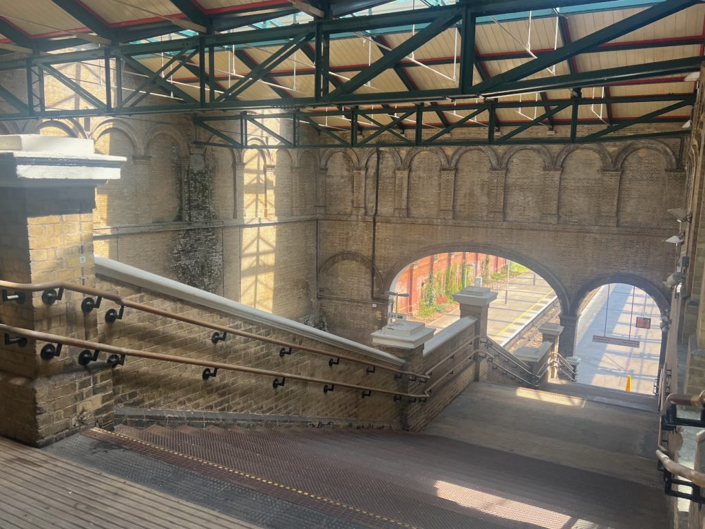 In a brick train shed, wide steps lead down from where the photograph stands towards the right, onto a platform that is visible through an arch in the far wall. A matching arch on the other side of the wall is blank, while a wide arch between the two reveals a second platform, between a railway track and a bright red wall. Above those arches are blank ones that surround the walls. The roof beams are cream-pained wood, held up by red and dark green metal beams. Light from a skylight forms shadows matching the beams on the side wall.