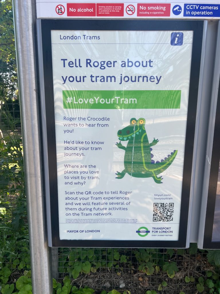 A poster in a frame, which reads: "London Trams. Tell Roger about your tram journey. #LoveYourTram. Roger the Croocdile wants to hear from you! He'd like to know about your tram journeys. Where are the places you love to visit by tram, and why? Scan the QR code to tell Roger about your Tram experiences and we will feature several of them during future activities on the Tram network. MAYOR OF LONDON; TRANSPORT FOR LONDON, EVERY JOURNEY MATTERS." The poster also contains a cartoon crocodile, whose dark-gren skin has a light-green line notched with arrows running across it, representing the trams on the Tube map. The poster is in a standard frame, with foliage behind it.