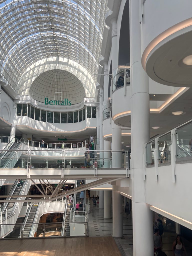 A view along a shopping centre galleria, with four floors in the main mall plus a fifth overlooking the mall from the department store at the end, Bentalls. The view is from the first floor. The walls are support columns are painted white, with cream-coloured trim lights along the bottom of each balcony. The ceiling is a semi circular arch, entirely glazed, stretching over the width of the mall; where it meets the curved windowed wall of Bentalls, they meet in a sphere. It's probably postmodern or something.