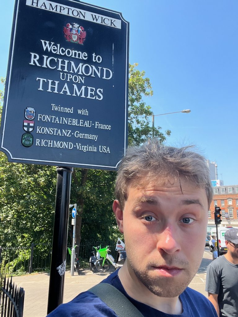 A selfie of the blog's author, wearing a blue t-shirt, with a vaguely shocked expression on his face. He stands below a black rectangular sign, with quarter-circles cut of of the corners. At the top of the sign is a white box, with "HAMPTON WICK" inside it. Below is the borough coat of arms, then, in white text: "Welcome to RICHMOND upon THAMES. Twinned with: FONTAINEBLEAU, France; KONSTANZ, Germany; RICHMOND, Virginia USA." Each twin town is listed to the right of a small version of its own coat of arms (or, in the case of Richmond, Virginia, a picture of an equestrian statue surrounded by the name of the city). In the background is a busy street, with foliage to the left and a red-brick building to the right. The sky is—yeah, you guessed it—a cloudless blue.
